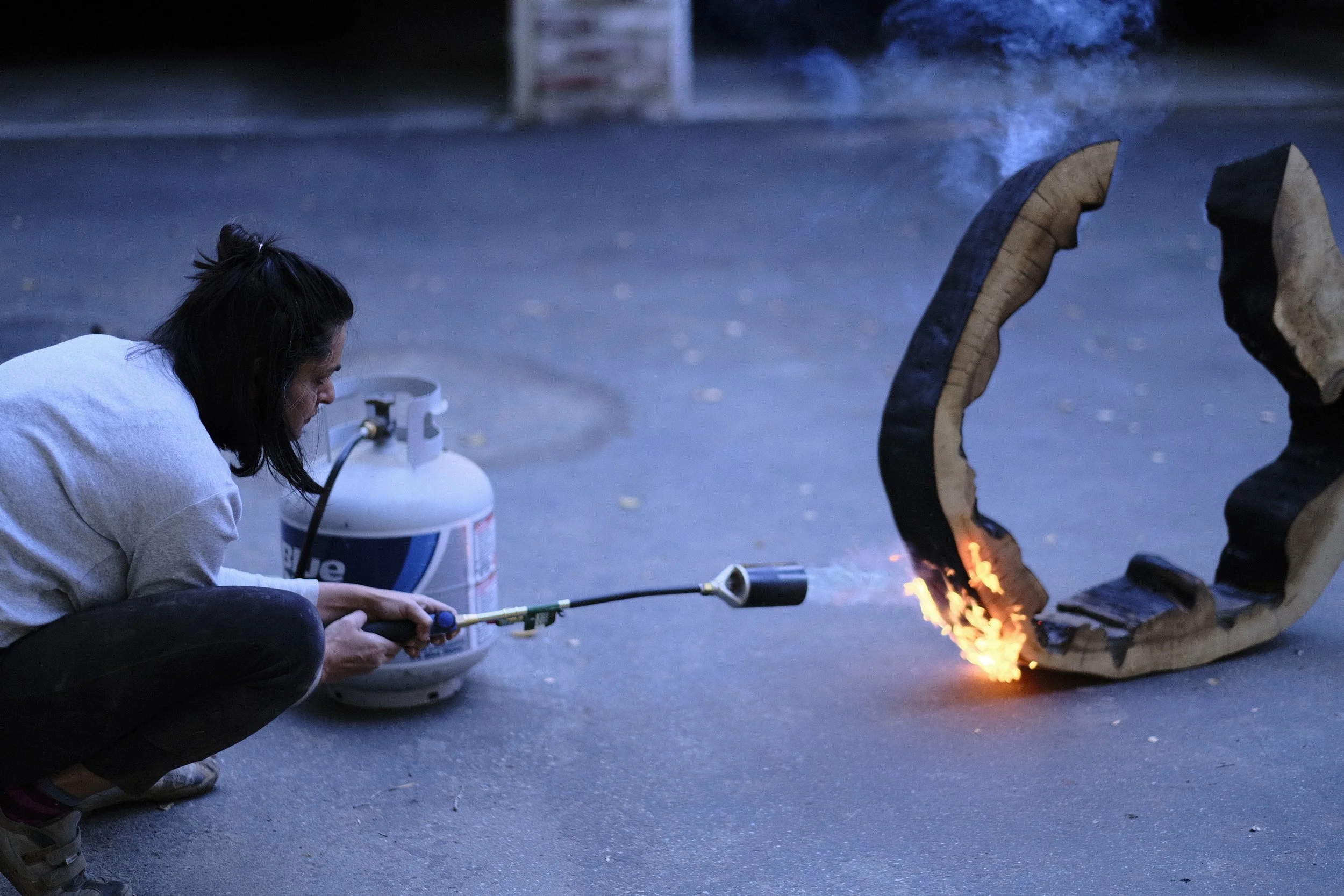 The artist crouched at dusk with a propane torch, flame licking a carved crescent of redwood