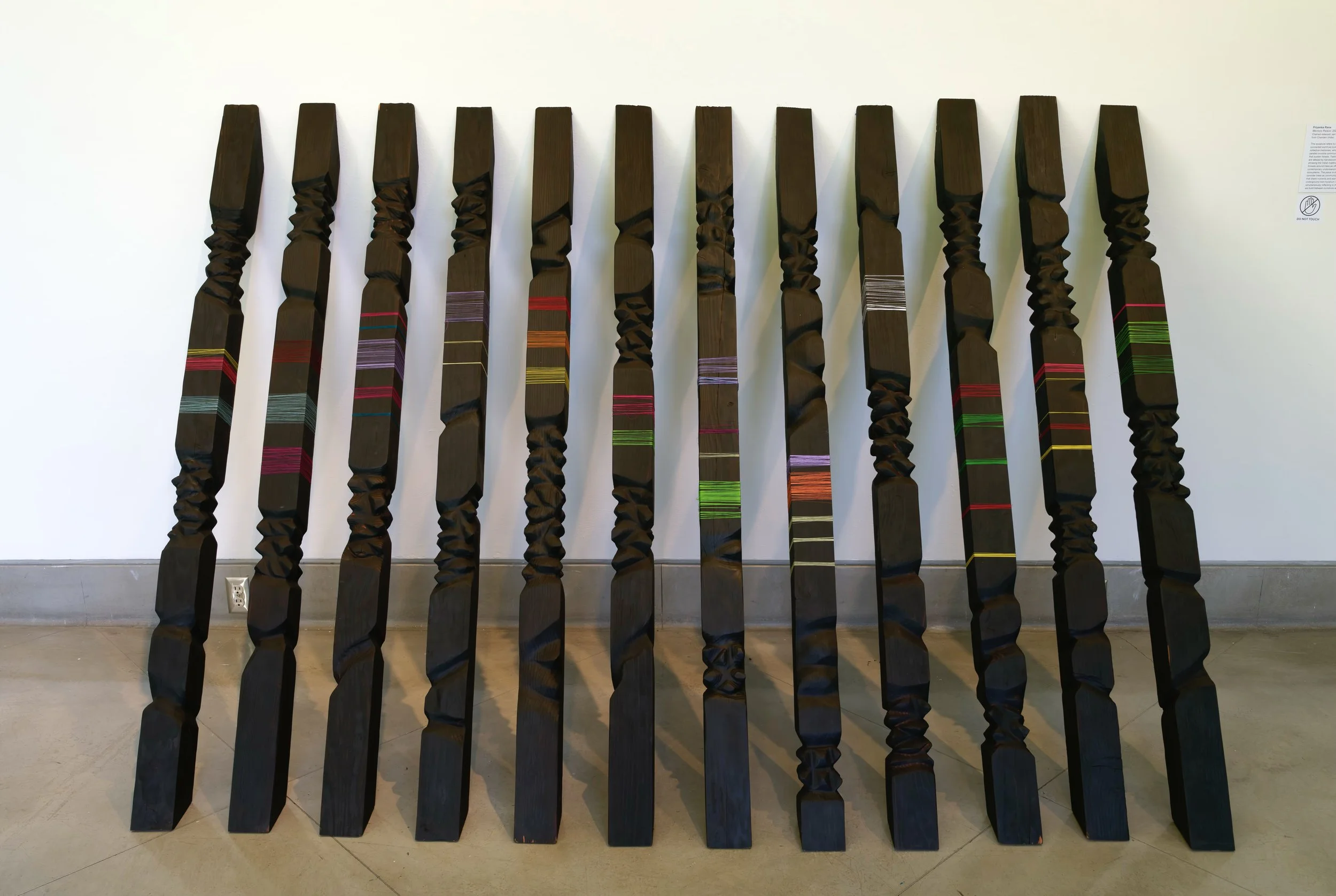Memory Palace — twelve carved and charred redwood pillars in a row, interwoven with bands of colored handwoven thread