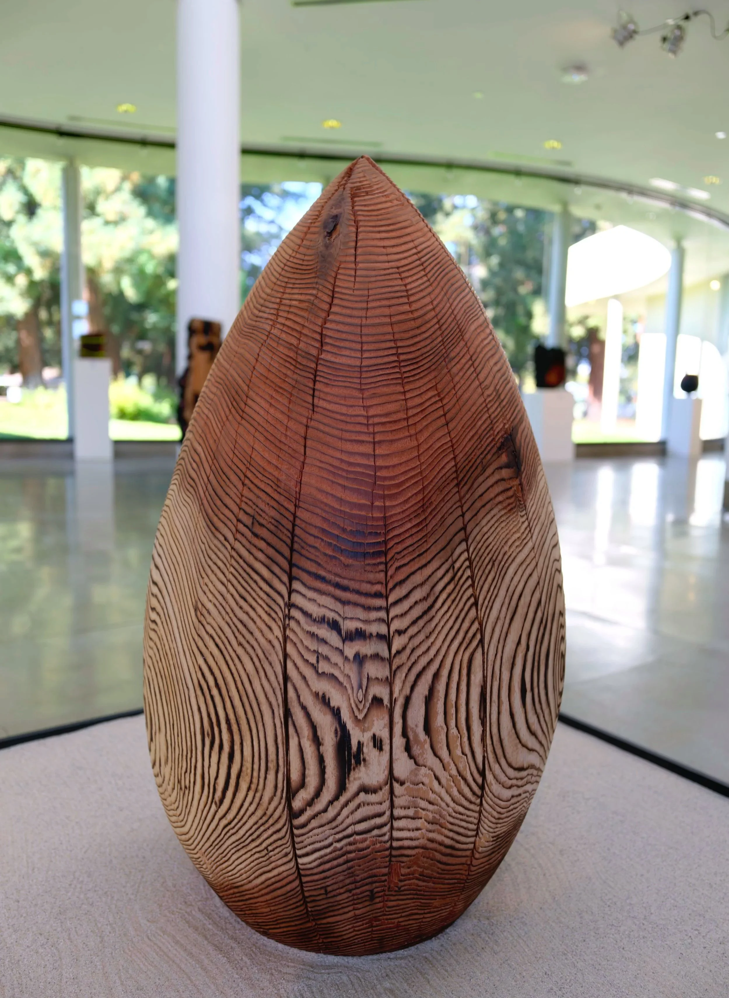 Monsoon — a carved redwood teardrop with striped charred grain, resting in a bed of sand in a bright gallery