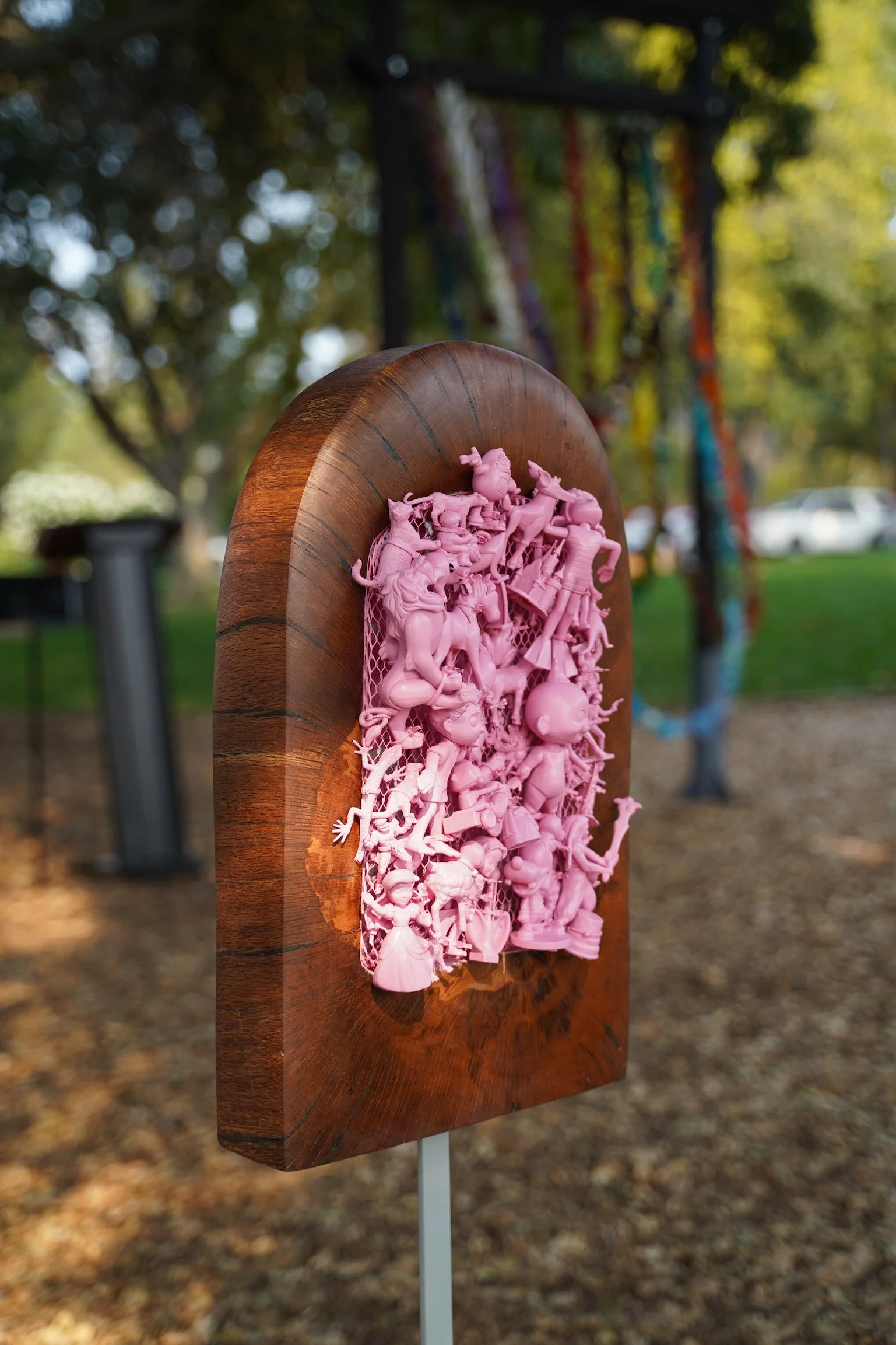 What’s At Play — a carved redwood arch holding a mass of pink spray-painted toys, installed in a playground