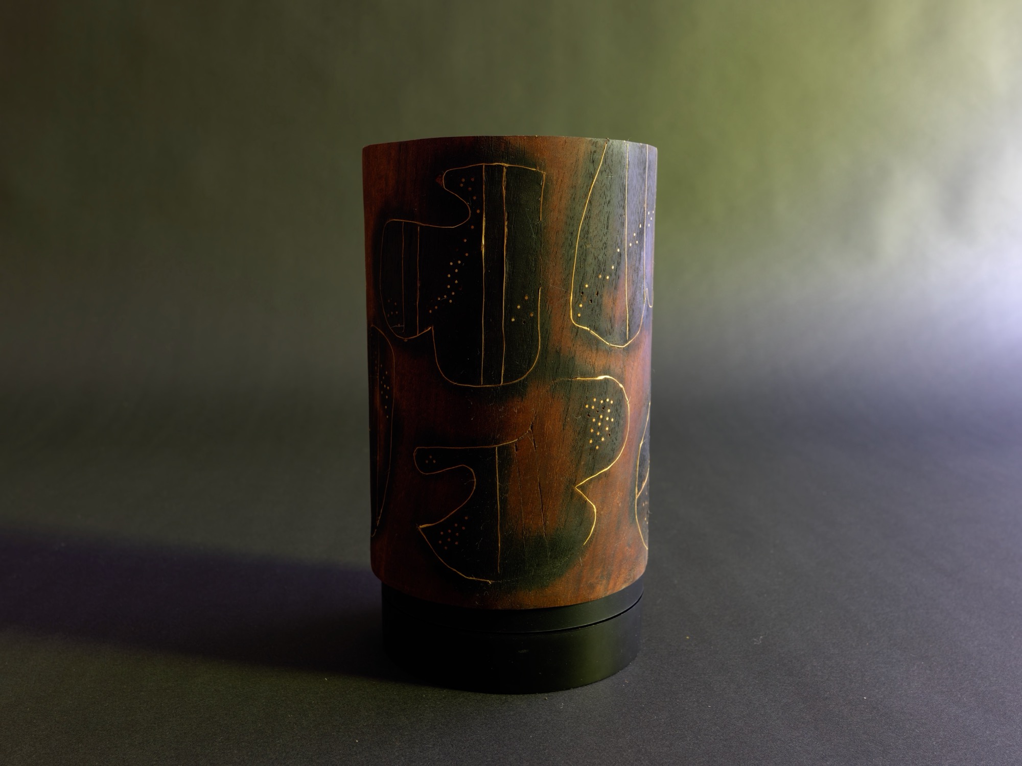 Dream — a carved cylindrical wood form incised with abstract figures, warm red-brown surface against a moss-green studio backdrop