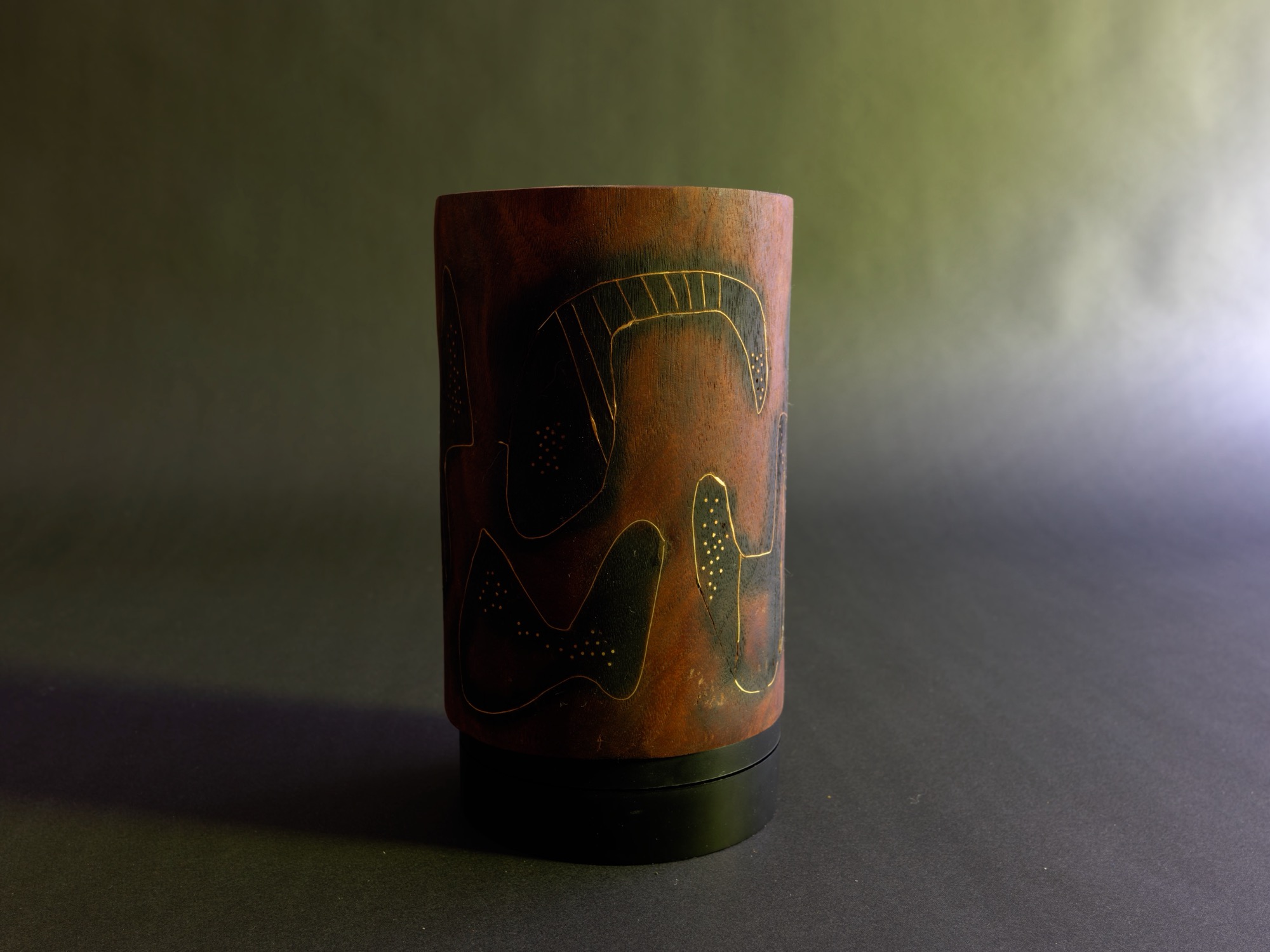Dream — a carved cylindrical wooden form incised with abstract figures in warm red and deep green, spotlit against a dark green studio backdrop