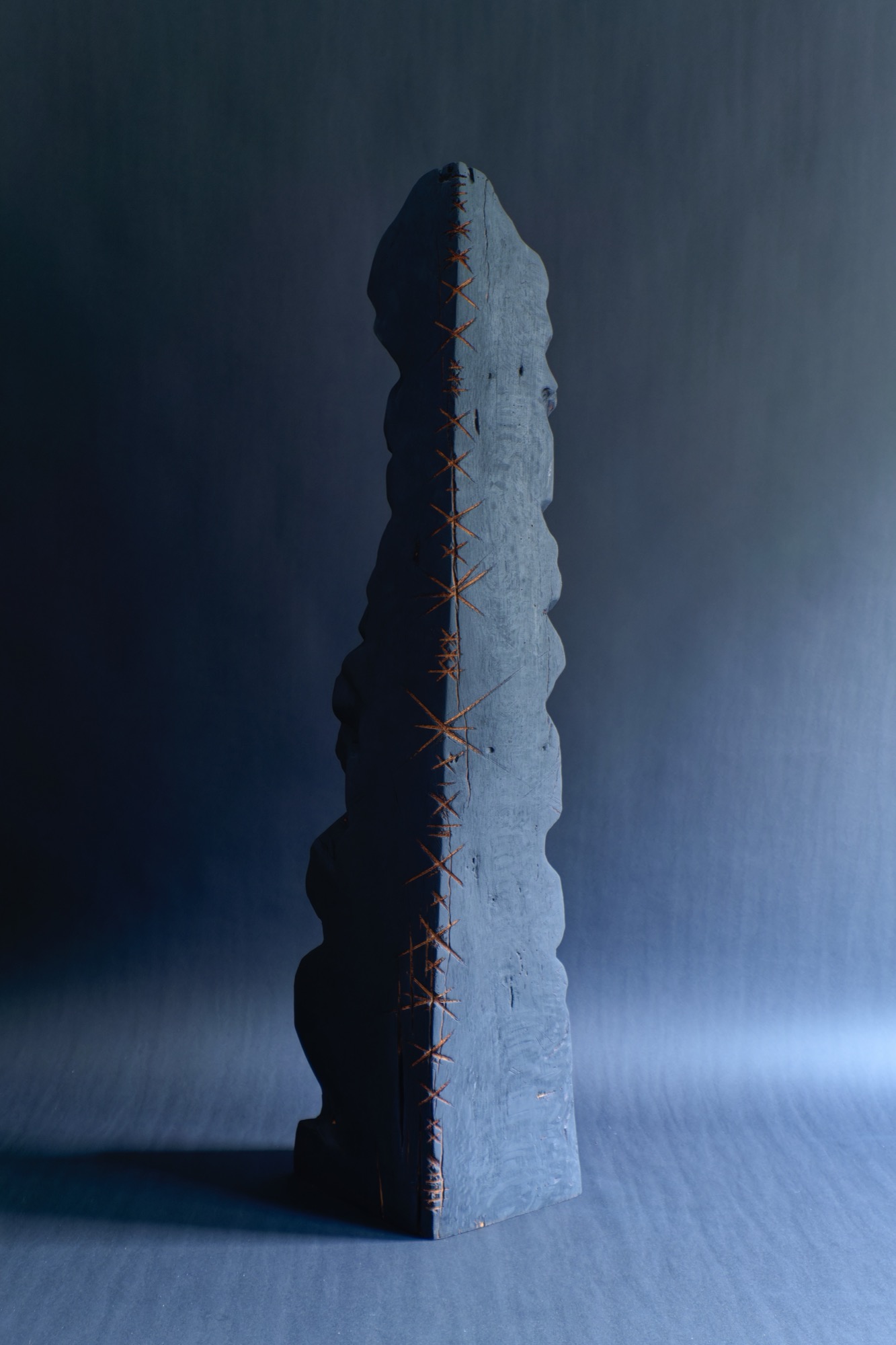 Healing 3 — a tall charred monolith stitched along its spine with copper thread, against a blue-grey wall